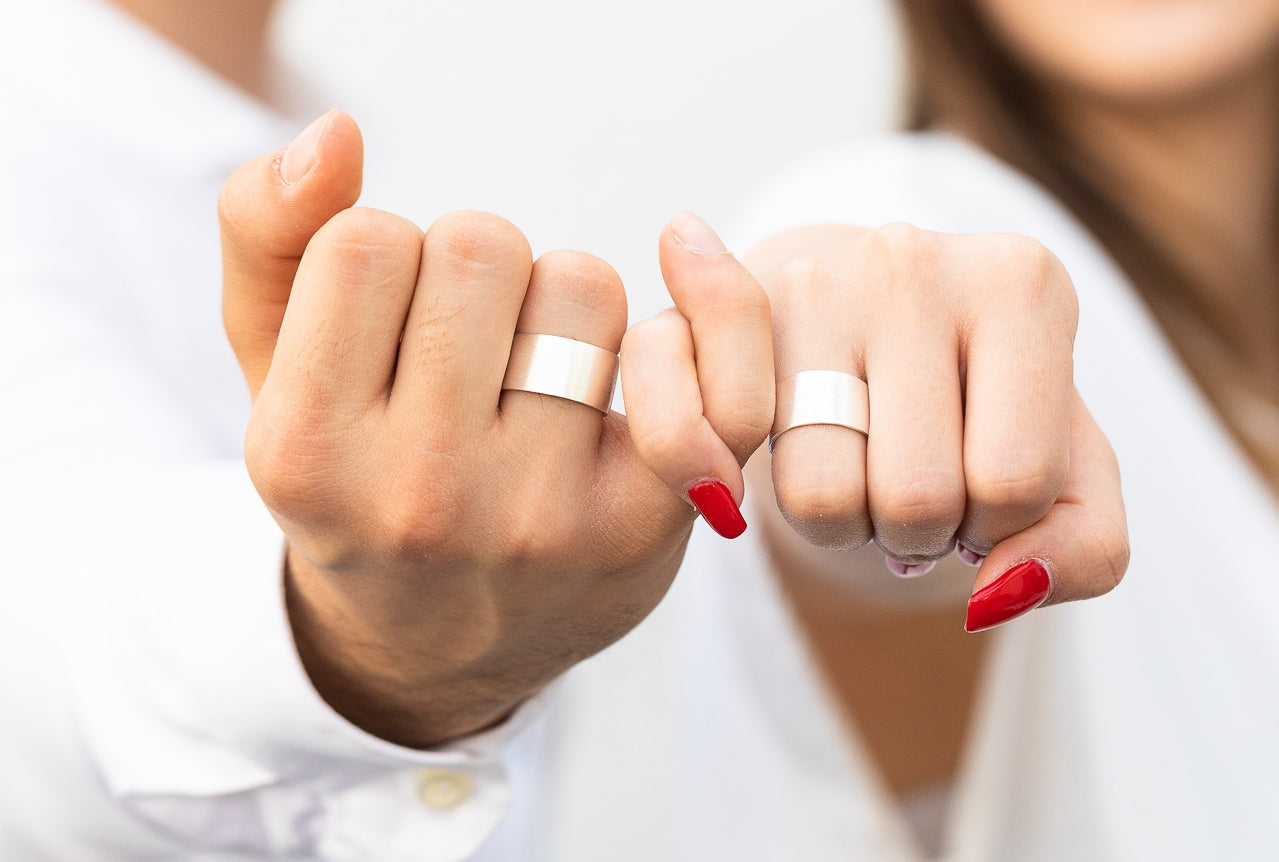 Zwei klassiche Bandringe aus Silber für Damen und Herren mit breiter Ringschiene und mattierter Oberfläche, getragen als Partnerringe an einer Damenhand und einer Herrenhand.