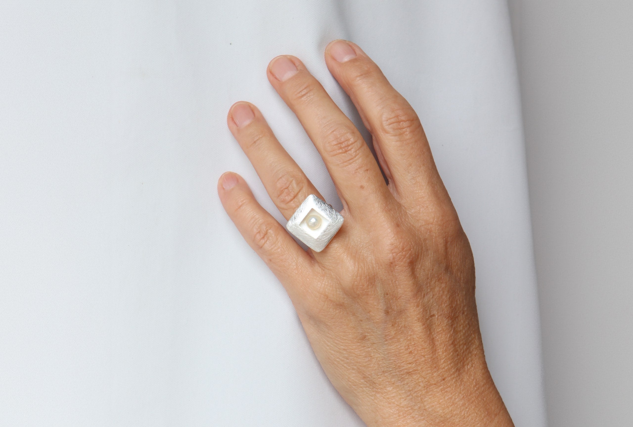 Ring aus Silber mit einer Perle getragen an einer Damenhand. Die Perle sitzt mittig in einem quadratischen Zierelement und die Ringschiene ist klassisch gearbeitet.