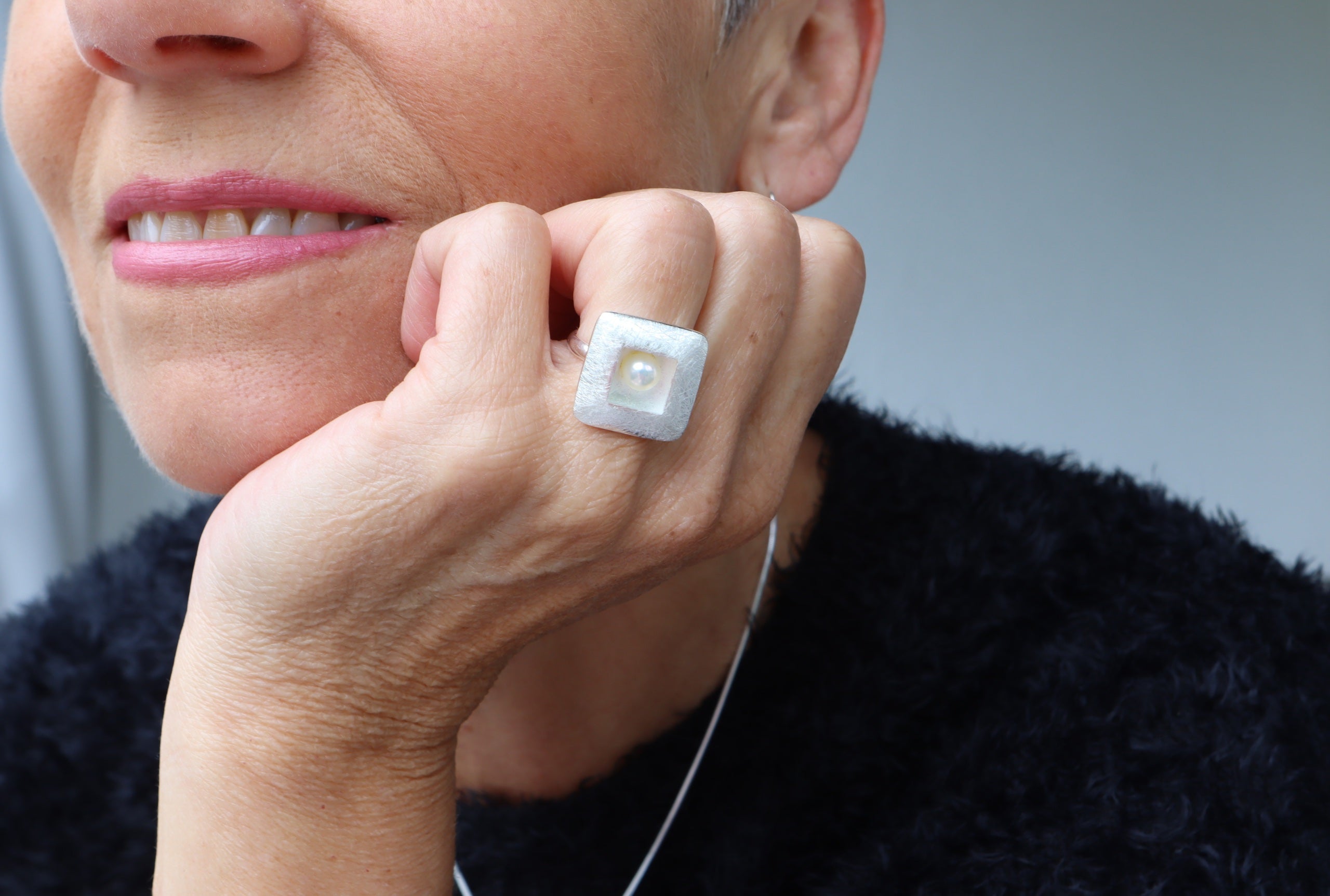 Ring aus Silber mit einer Perle getragen an einer Damenhand. Die Perle sitzt mittig in einem quadratischen Zierelement und die Ringschiene ist klassisch gearbeitet.