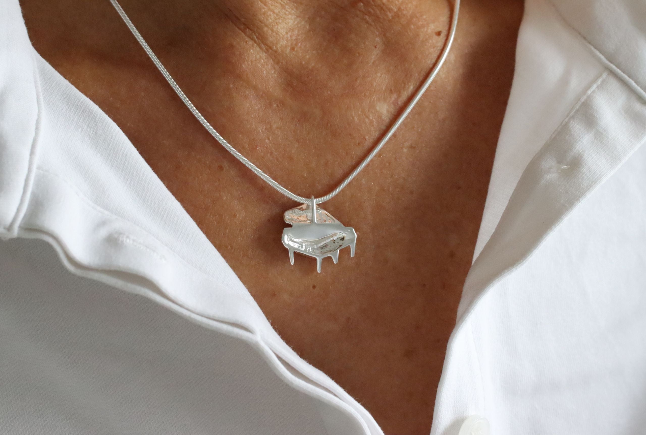 Ein Anhänger aus Silber, der ein fein herausgearbeitetes Klavier darstellt und eine glänzende Oberfläche hat, getragen mit einer Schlangenkette.