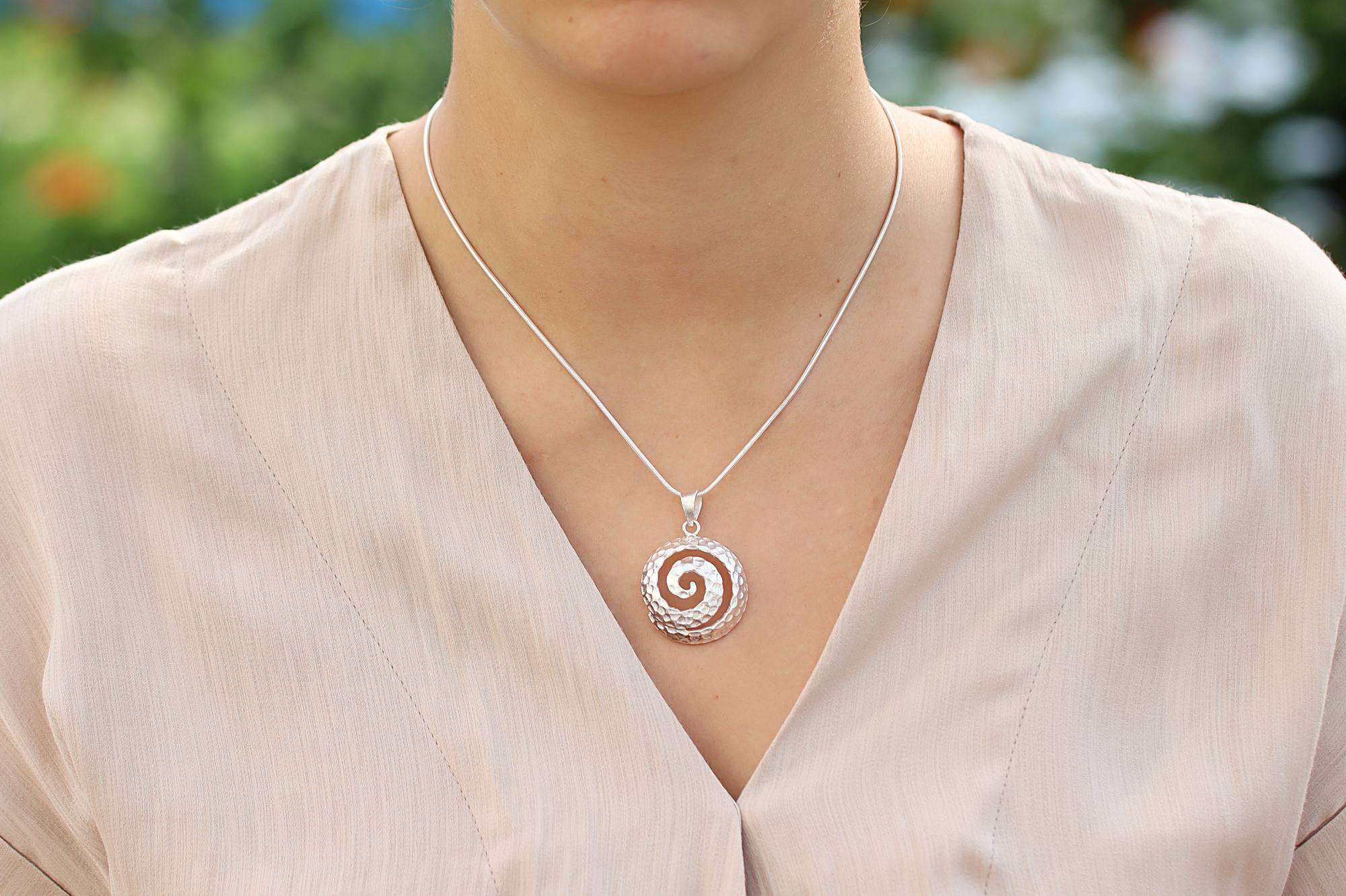 Geschmiedeter Anhänger aus Silber für Damen in Form einer Spirale mit glänzend gehämmerter Oberfläche, getragen mit einer Schlangenkette an einem Modell. 