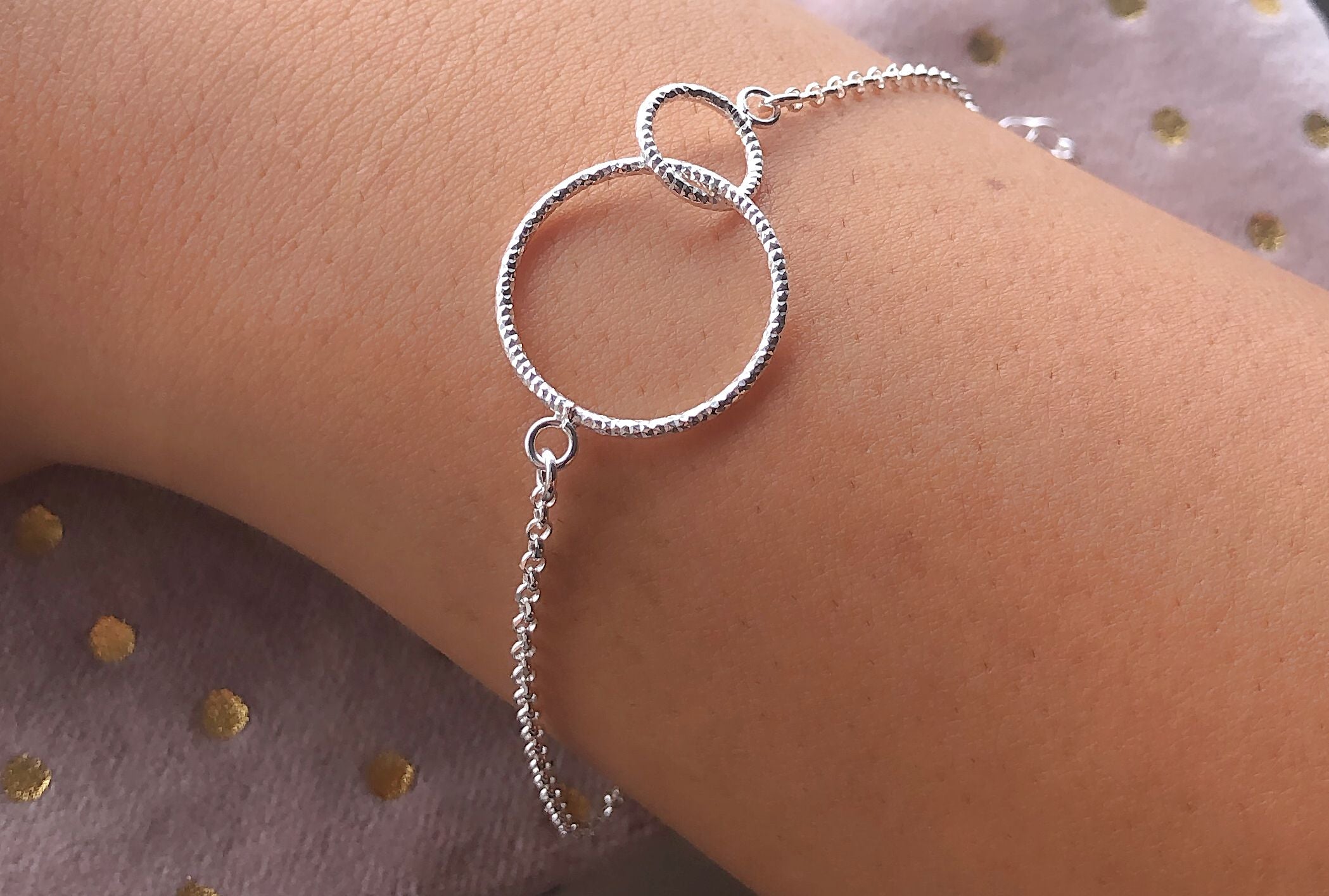 Ein Armband aus Silber aus einer Ankerkette mit zwei ineinander greifenden Kreisen, getragen am Handgelenk einer Dame.