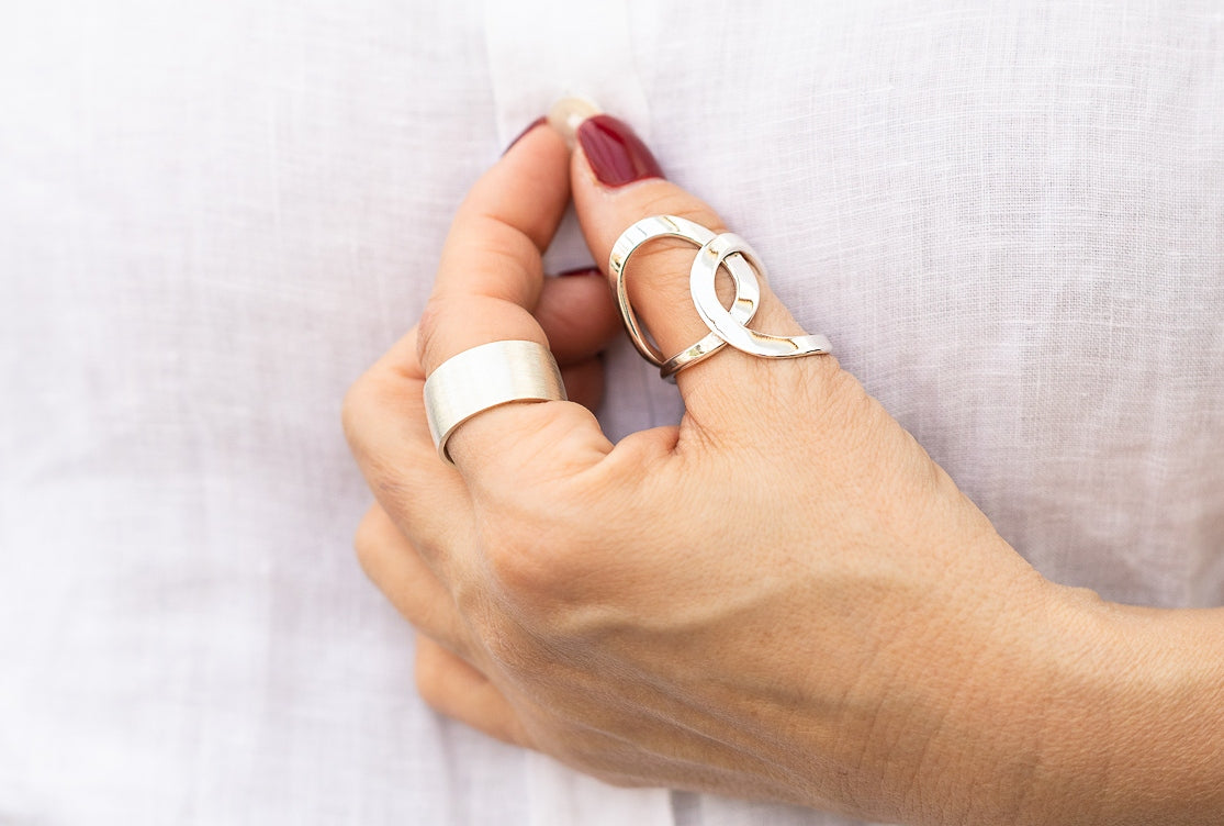 Geschmiedeter Damenring aus zwei Kreisen, die kunstvoll ineinander verschlungen sind, getragen am Daumen einer Damenhand. Der Ring ist geschmiedet und hat eine glänzende Oberfläche.